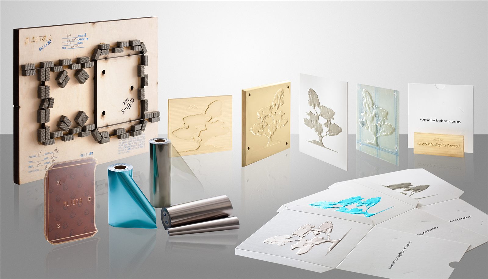 Folding carton tooling: a die-cutting forme with rubber ejection, brass foil-stamping and embossing dies, foil rolls, and finished embossed and foil-stamped packaging samples.
